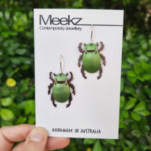 Load image into Gallery viewer, Green Christmas Beetle earrings made from car vinyl, recycled copper, sterling‑silver hooks, and stainless‑steel jump rings, shown displayed on a branded jewellery card. The beetle’s metallic green body catches the light with a vivid sheen, while the darker legs and antennae add contrast. The clean white card and outdoor greenery frame the piece, highlighting its shine, texture, and detailed insect‑inspired craftsmanship.