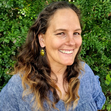 Load image into Gallery viewer, Gold King Christmas Beetle earring made from reflective gold car vinyl, recycled copper, a sterling‑silver hook, and stainless‑steel jump rings, modelled by Jemica. The beetle’s domed gold body is intensely reflective, catching bright highlights as it hangs from the ear, while the darker metal legs and antennae add crisp contrast. The outdoor greenery softens the background, drawing focus to the earring’s shine, texture, and detailed insect‑inspired form.