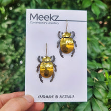 Load image into Gallery viewer, Gold King Christmas Beetle earring made from reflective gold car vinyl, recycled copper, a sterling‑silver hook, and stainless‑steel jump rings, shown displayed on a branded jewellery card. The beetle’s highly reflective gold body catches bright highlights, contrasted by dark metal legs and antennae. The card is held outdoors against soft green foliage, emphasizing the earring’s shine, texture, and detailed insect‑inspired form.