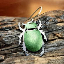 Load image into Gallery viewer, Green Christmas Beetle earring made from car vinyl, recycled copper, a sterling‑silver hook, and stainless‑steel jump rings, shown resting on textured wood. The beetle’s metallic green body catches the light with a vivid sheen, while the copper legs and antennae add crisp contrast. The close, natural setting highlights the earring’s shine, segmented form, and detailed insect‑inspired craftsmanship.