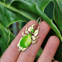 Load image into Gallery viewer, Leaf insect earrings - the image shows a leaf mimicking insect earring made from lime‑green car vinyl with recycled brass legs and detailing, a sterling‑silver hook, and hypoallergenic stainless‑steel jump rings, shown held in a hand against large green leaves.