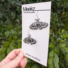 Load image into Gallery viewer, A different viewpoint of the Mistletoe Moth Earrings displayed on a packaging card, being held up in front of a green plant wall. They are made of copper with a black patina and have hand painted white zigzag patterns on the wings.