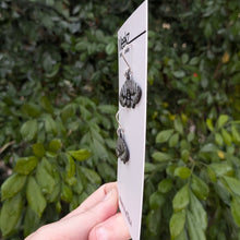 Load image into Gallery viewer, A different viewpoint of the Mistletoe Moth Earrings displayed on a packaging card, being held up in front of a green plant wall. They are made of copper with a black patina and have hand painted white zigzag patterns on the wings.