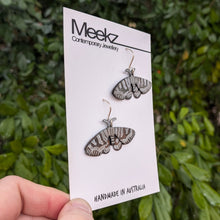 Load image into Gallery viewer, A different viewpoint of the Mistletoe Moth Earrings displayed on a packaging card, being held up in front of a green plant wall. They are made of copper with a black patina and have hand painted white zigzag patterns on the wings.