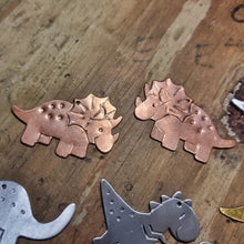 Load image into Gallery viewer, Copper Triceratops dinosaur-shaped earrings on a wooden jewellers bench surrounded by other dinosaurs