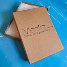 Load image into Gallery viewer, Meekz gift box made from kraft‑brown card, stamped with the Meekz Contemporary Jewellery logo on the lid. The box is shown partially open with soft white cushioning inside, highlighting the protective packaging the earrings come in. The blue surface beneath adds contrast, drawing attention to the clean branding and simple, eco‑minded presentation.