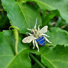 Load image into Gallery viewer, Blue‑banded bee earring made from blue car vinyl with recycled brass wings and body details, a sterling‑silver hook, and hypoallergenic stainless‑steel jump rings, displayed on green leaves with vine details.
