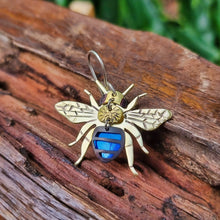 Load image into Gallery viewer, Blue banded bee earring made from blue car vinyl with recycled brass wings and body details, a sterling‑silver hook, and hypoallergenic stainless‑steel jump rings, displayed on textured wood with soft green foliage in the background.
