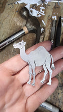 Load image into Gallery viewer, Hand holding a metal cutout of a camel with tools and paper shards in the background
