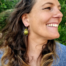 Load image into Gallery viewer, Gold King Christmas Beetle earring made from gold car vinyl, recycled copper, a sterling‑silver hook, and stainless‑steel jump rings, shown worn on a model. The metallic gold beetle hangs just below the ear, its reflective body catching natural outdoor light, with copper legs and antennae adding crisp detail. Soft greenery in the background frames the earring, highlighting its warm shine and sculptural, insect-inspired form.

