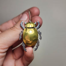 Load image into Gallery viewer, Gold King Christmas Beetle earring made from reflective gold car vinyl, recycled copper, a sterling‑silver hook, and stainless‑steel jump rings, shown  close‑up held between fingers. The beetle’s domed gold body is intensely reflective, catching bright highlights across its surface, while the darker metal legs and antennae create sharp contrast. The tight framing emphasizes the shine, texture, and detailed insect‑inspired craftsmanship.

