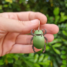 Load image into Gallery viewer, Green Christmas Beetle earring made from car vinyl, recycled copper, a sterling‑silver hook, and stainless‑steel jump rings, shown held between fingers in a close‑up. The beetle’s metallic green body catches natural light with a vivid sheen, while the darker legs and antennae add contrast. The tight framing highlights the earring’s shine, texture, and detailed insect‑inspired craftsmanship.
