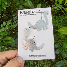Load image into Gallery viewer, Diplodocus (Long Neck) Dinosaur-shaped earrings on a Meekz Packaging card held in front of green foliage
