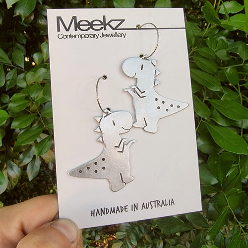 Aluminium dinosaur shaped earrings on a 'Meekz Contemporary Jewellery' card with green leaves in the background