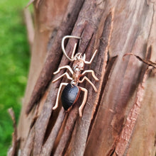 Load image into Gallery viewer, Close‑up of a meat‑ant–inspired earring made from black car vinyl with recycled copper body segments, a sterling‑silver hook, and a hypoallergenic stainless‑steel jump rings, shown hanging on tree bark with soft green grass in the background.
