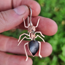 Load image into Gallery viewer, Close‑up of a meat‑ant–inspired earring made from black car vinyl with recycled copper body segments, a sterling‑silver hook, and a hypoallergenic stainless‑steel jump rings, held in a hand against a soft green leafy background.
