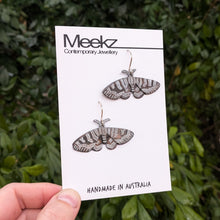 Load image into Gallery viewer, Mistletoe Moth Earrings displayed on a packaging card, being held  up in front of a green plant wall. They are made of copper with a black patina and have hand painted white zigzag patterns on the wings.
