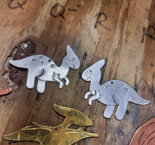 Load image into Gallery viewer, Aluminium Parasaurolophus dinosaur earrings on a wooden jewellers bench surrounded by other dinosaurs 
