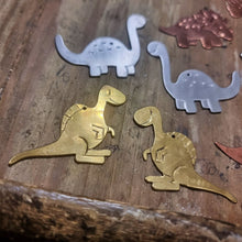 Load image into Gallery viewer, Brass Spinosaurus dinosaur-shaped earrings on a wooden jewellers bench surrounded by other dinosaurs
