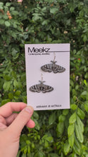 Load and play video in Gallery viewer, A video of the Mistletoe Moth Earrings displayed on a packaging card, being held  up in front of a green plant wall and moving side to side. They are made of copper with a black patina and have hand painted white zigzag patterns on the wings.  
