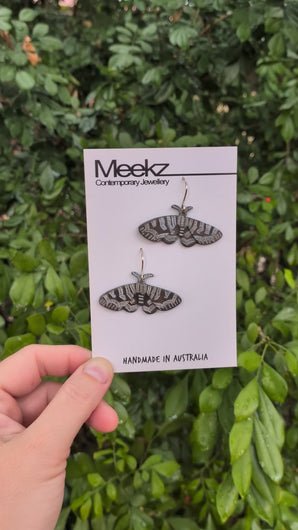 A video of the Mistletoe Moth Earrings displayed on a packaging card, being held  up in front of a green plant wall and moving side to side. They are made of copper with a black patina and have hand painted white zigzag patterns on the wings.  